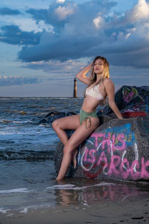 A beachwear model poses on graffiti-covered rocks at sunset with a lighthouse visible in the background by the ocean.