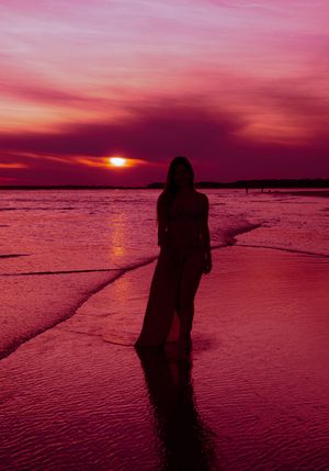A silhouette stands on a beach during a vibrant pink and purple sunset over calm ocean waters.