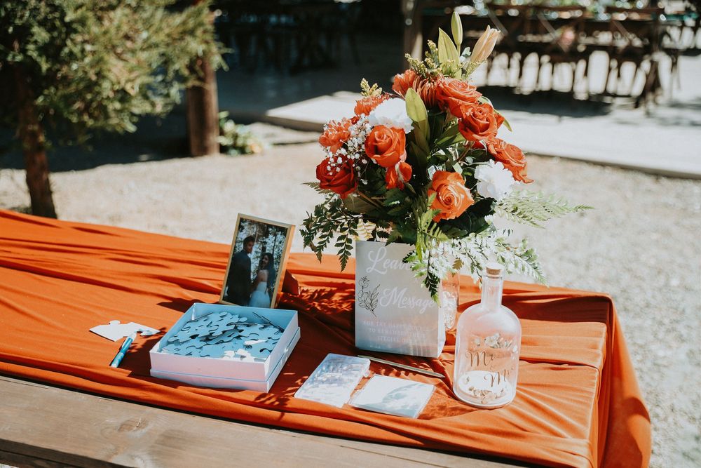 Orange floral centerpiece with coral roses sits on outdoor table with orange tablecloth and mason jar decor.
