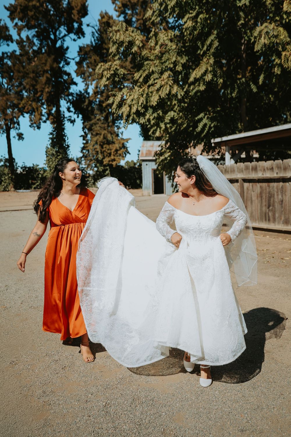 A bride in a white wedding gown and veil walks outdoors with a bridesmaid wearing a bright orange dress.