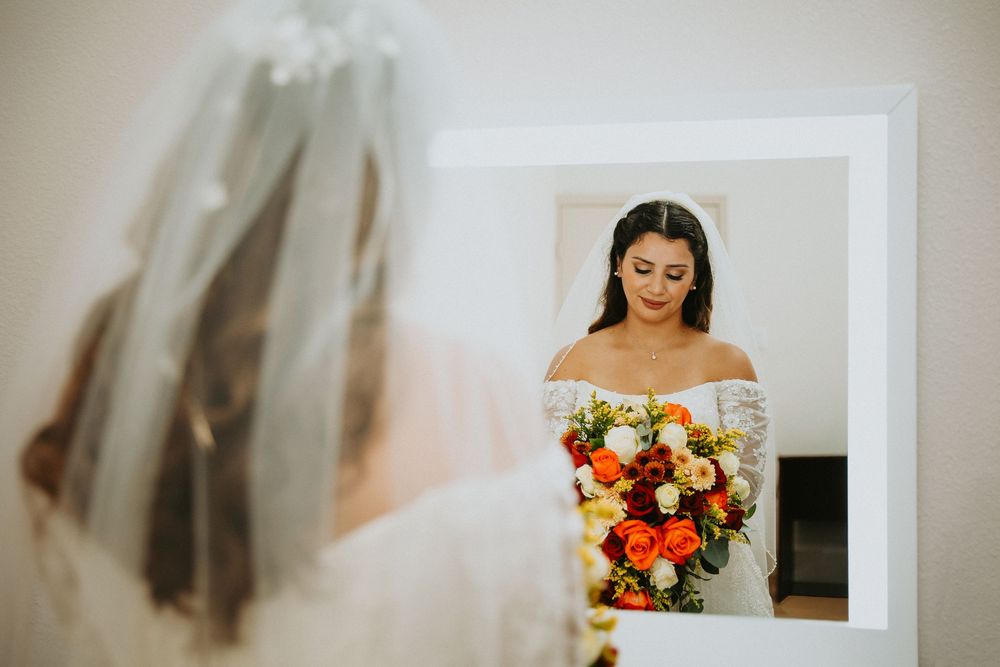 A bride in a white wedding dress holds a vibrant autumn-colored bouquet while getting ready in a sunlit room.