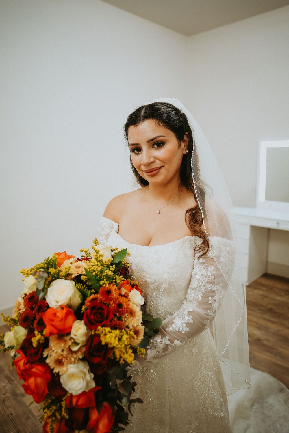 A bride in a white lace gown holds a vibrant autumn-colored bouquet with red and orange flowers.