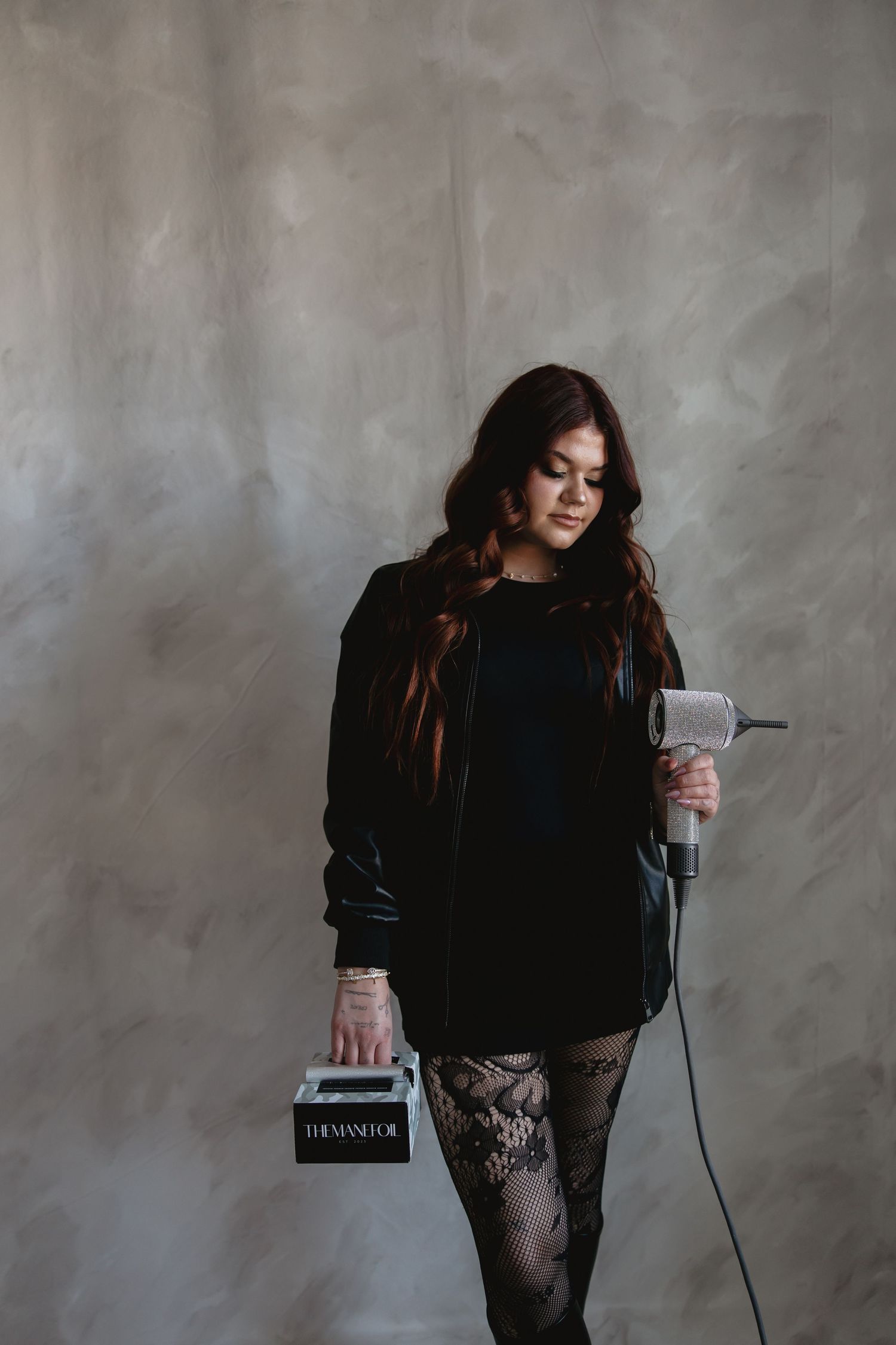 A person in a black sweater stands against a gray wall while holding a book and microphone.