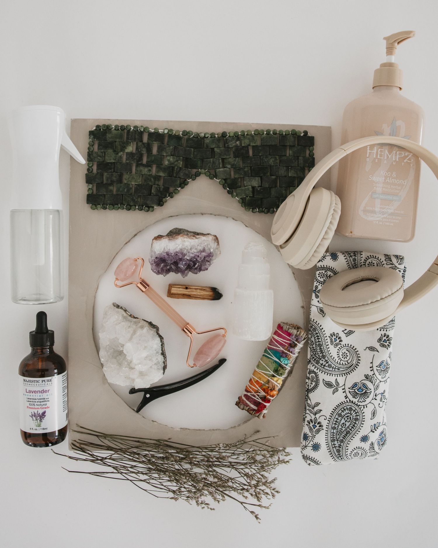 A flat lay of self-care items including a jade roller, amethyst crystal, essential oils, and skincare products on white surface.