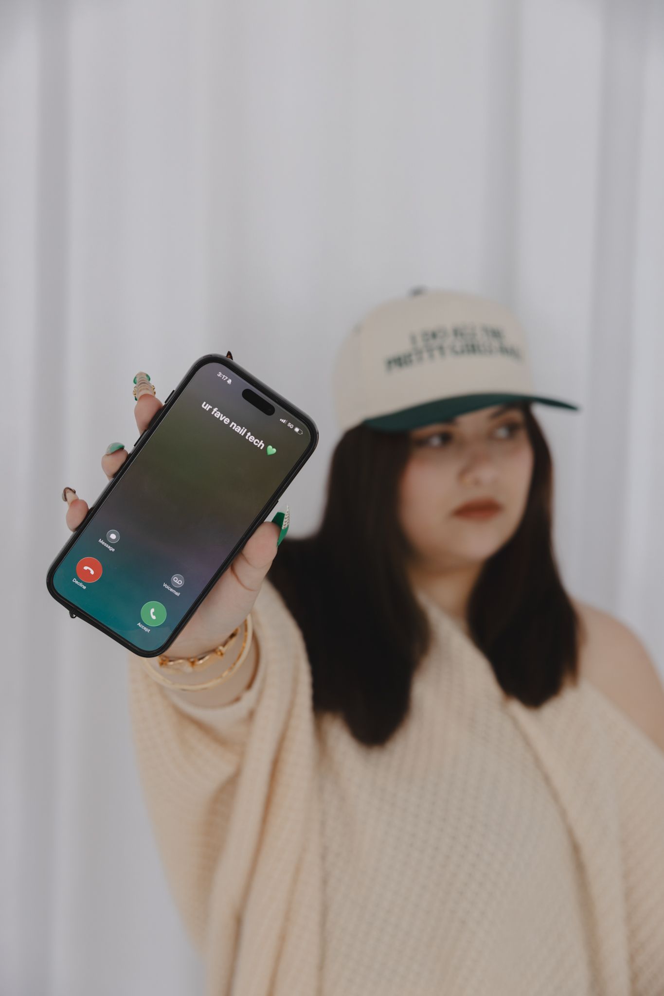 Woman in cream sweater and white cap holds iPhone showing incoming call screen toward camera.