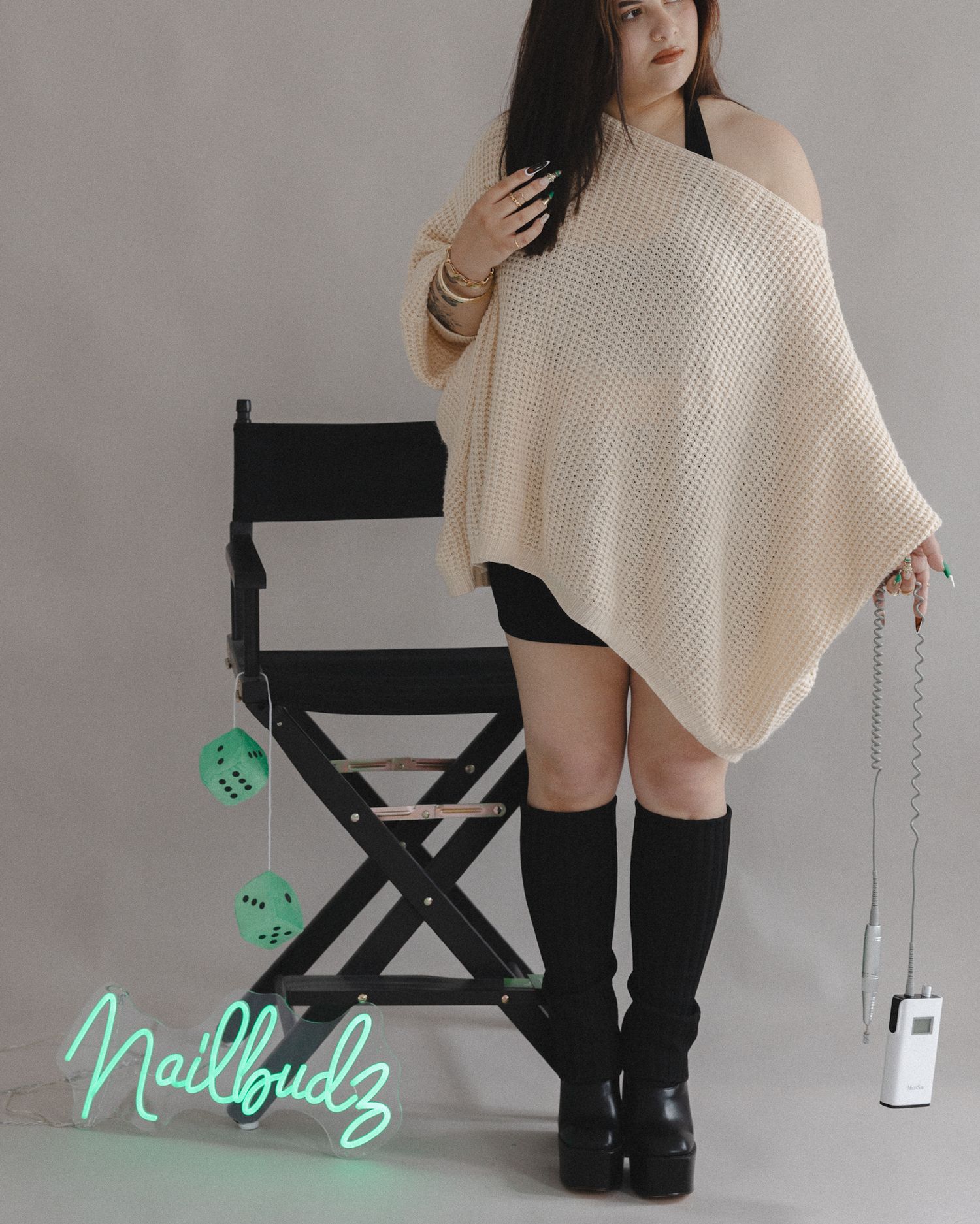 Woman in cream knit poncho, black shorts, and knee-high boots posing near a director's chair with Nailbudz neon sign.