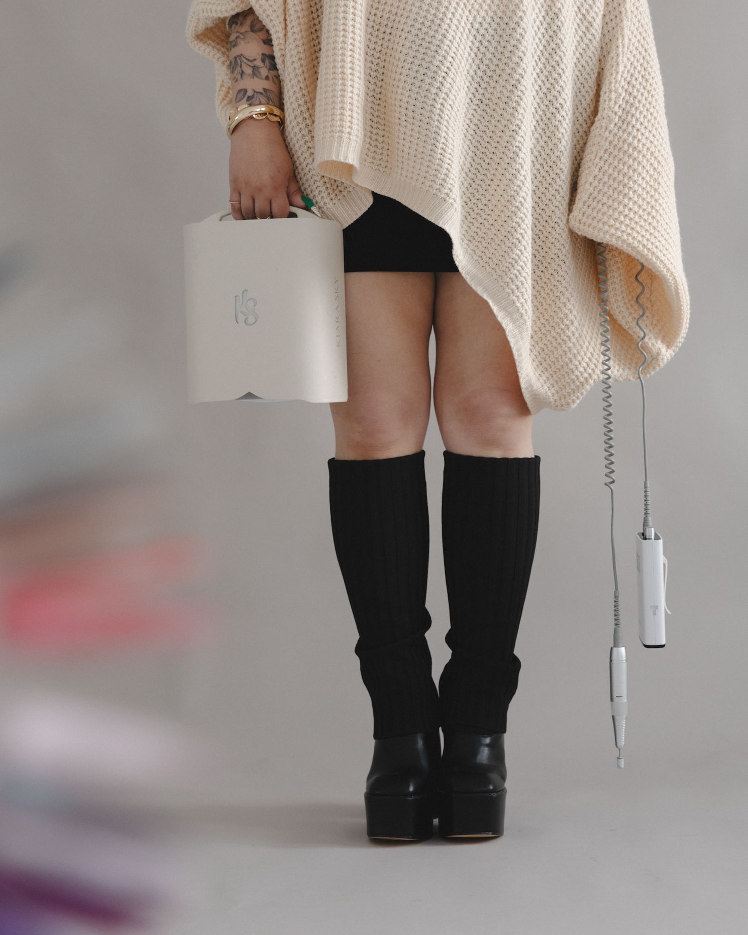Person wearing cream knit sweater, black shorts, knee-high boots, holding white handbag and hair tool.