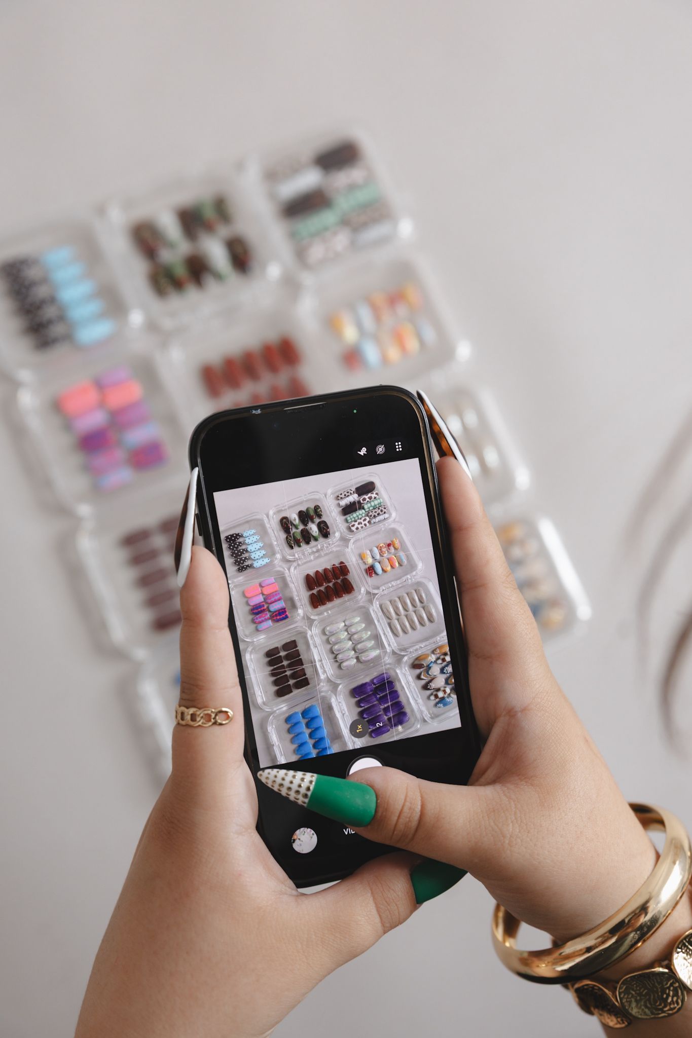 Woman with green nails and gold bracelet photographing colorful press-on nail sets displayed in clear packaging on white background.