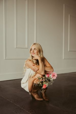 Blonde woman crouching in white dress and brown cowboy boots holding pink floral bouquet, smiling.