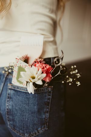 Close-up of jeans pocket holding hair scissors, comb, and fresh flowers tucked into waistband.