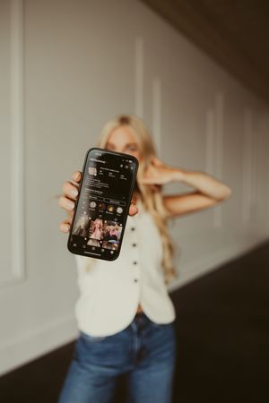 Blonde woman in white vest holding smartphone displaying Instagram profile toward camera.
