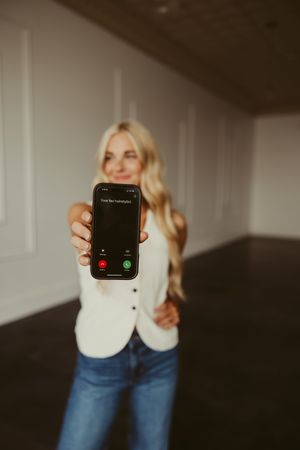 Blonde woman in white blazer and jeans holding up smartphone showing incoming call screen.