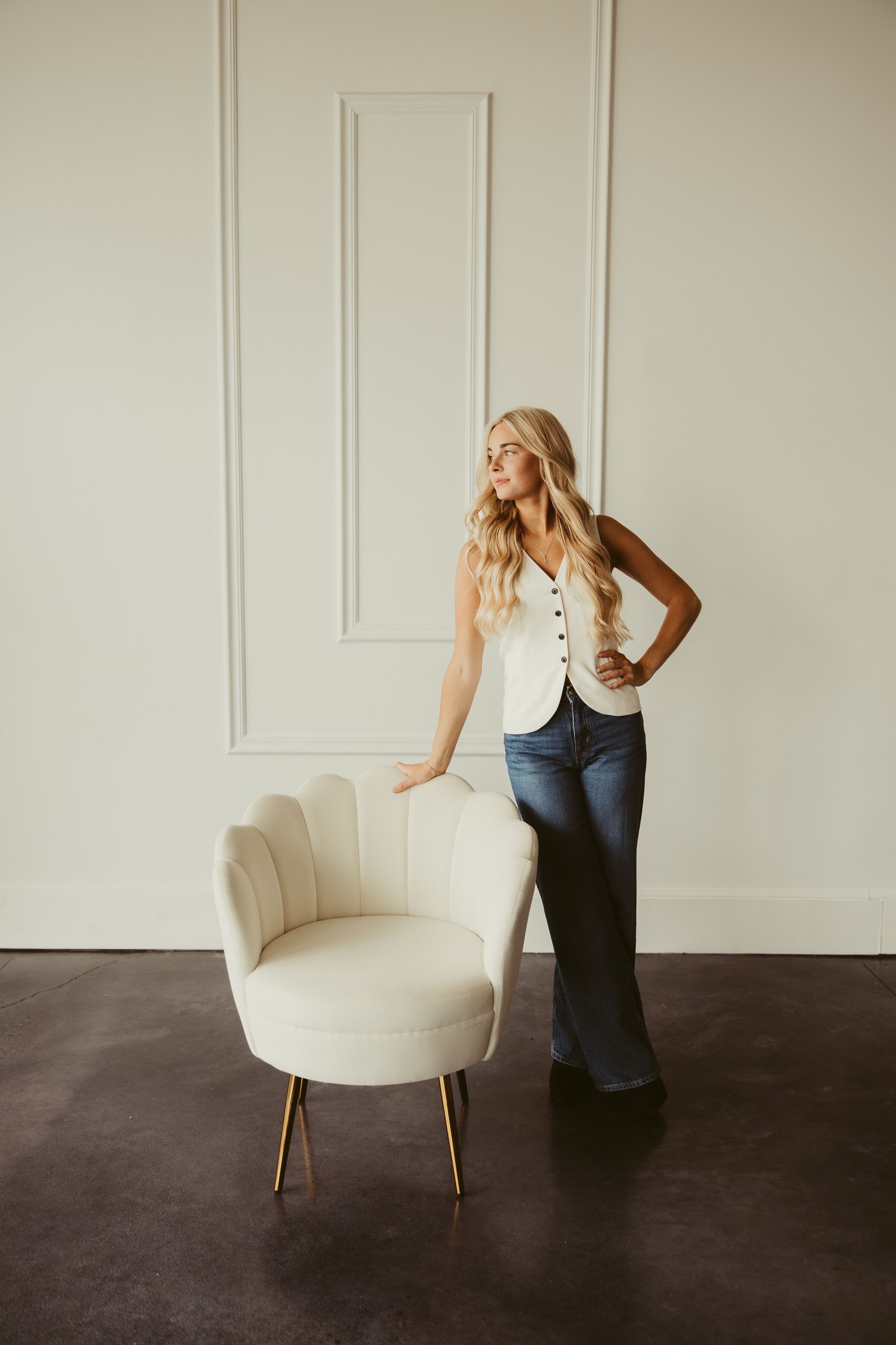 Blonde hairstylist in white vest and flare jeans posing beside cream velvet chair in white studio.