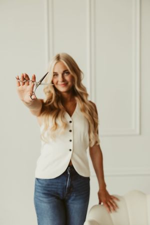 Smiling blonde hairstylist holding scissors toward camera in bright white paneled studio setting.