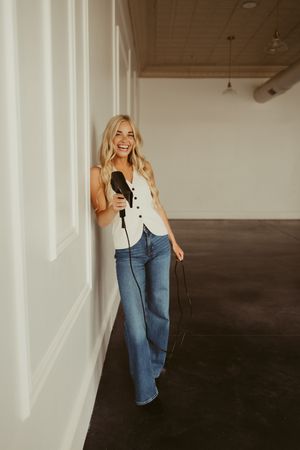Cheerful blonde hairstylist leaning against white wall holding hair dryer in modern salon studio.