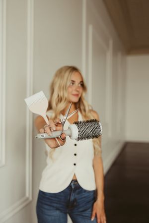 Blonde hairstylist holding scissors, round brush, and comb toward camera in white vest and jeans.