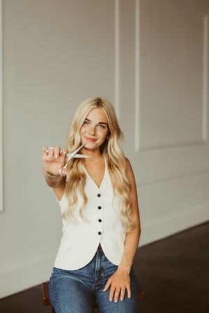 Smiling blonde woman in white button vest and jeans holding hair scissors toward camera.
