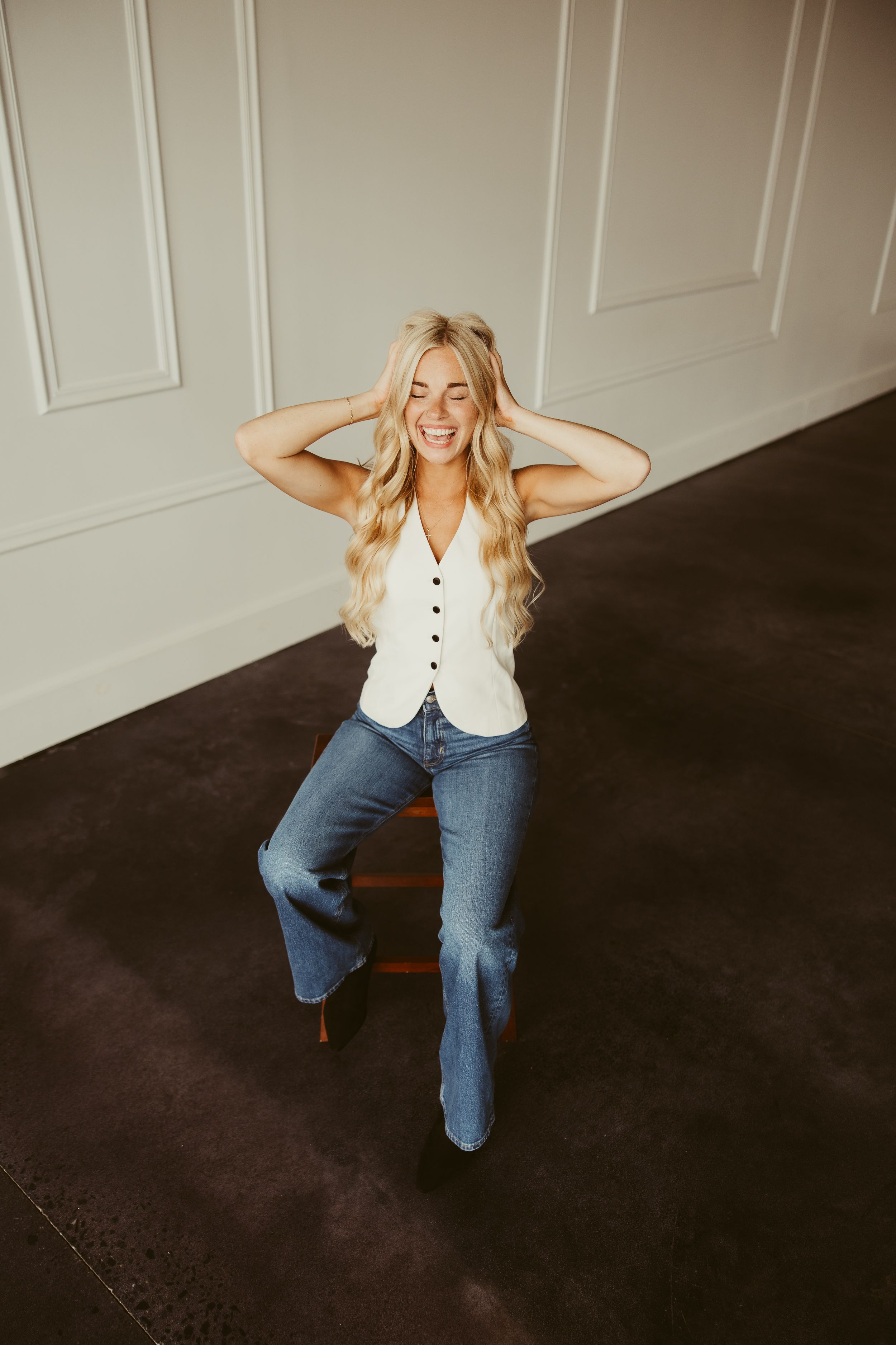 Joyful blonde woman sitting on wooden stool laughing with hands in hair, wearing white vest and jeans.