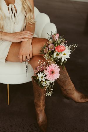 Close-up of blonde woman in white dress and cowboy boots with pink floral bouquet tucked in boot.