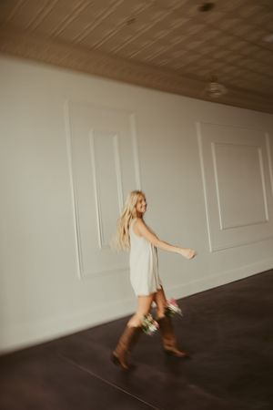 Motion-blurred photo of blonde woman in white dress and cowboy boots walking with floral bouquet.