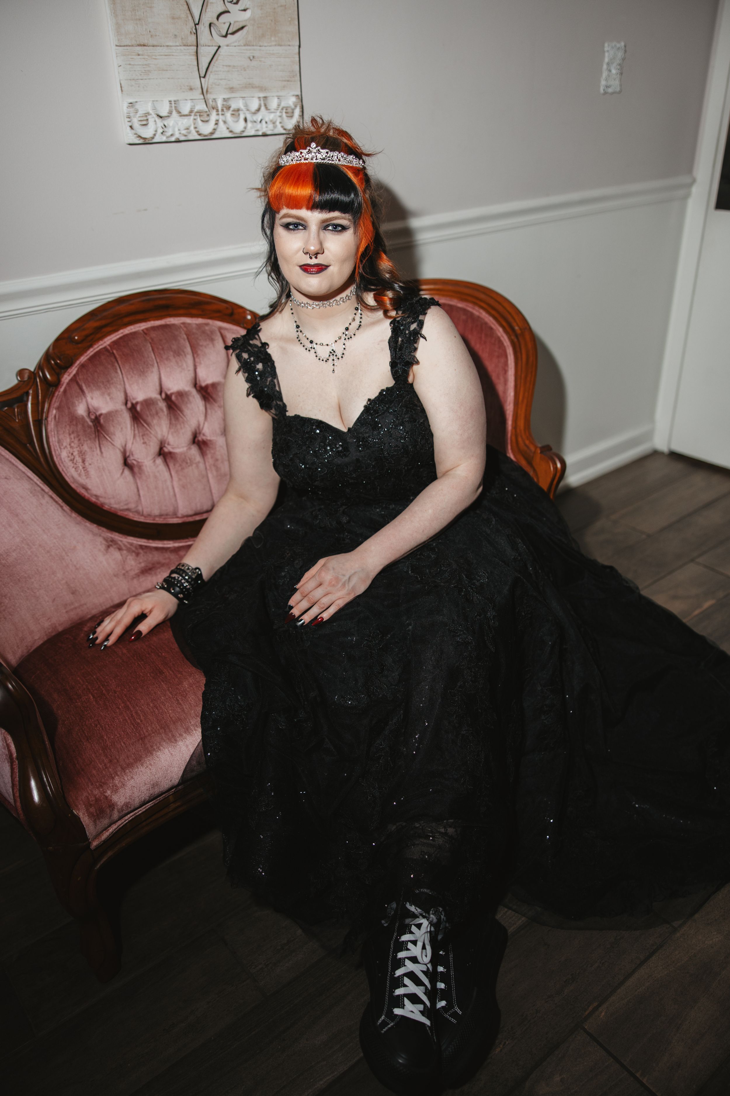 Gothic bride in black dress and orange headpiece lounges on vintage pink velvet couch.