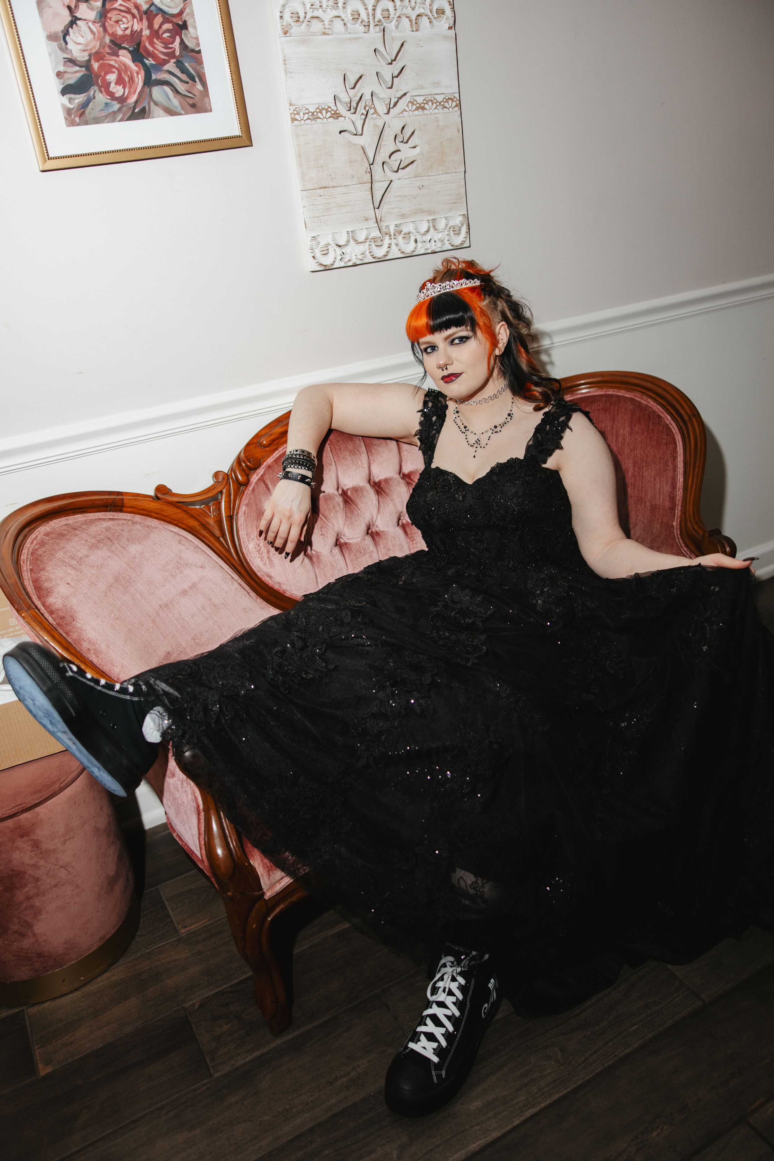 Alternative bride in black dress and combat boots reclines on pink velvet couch.
