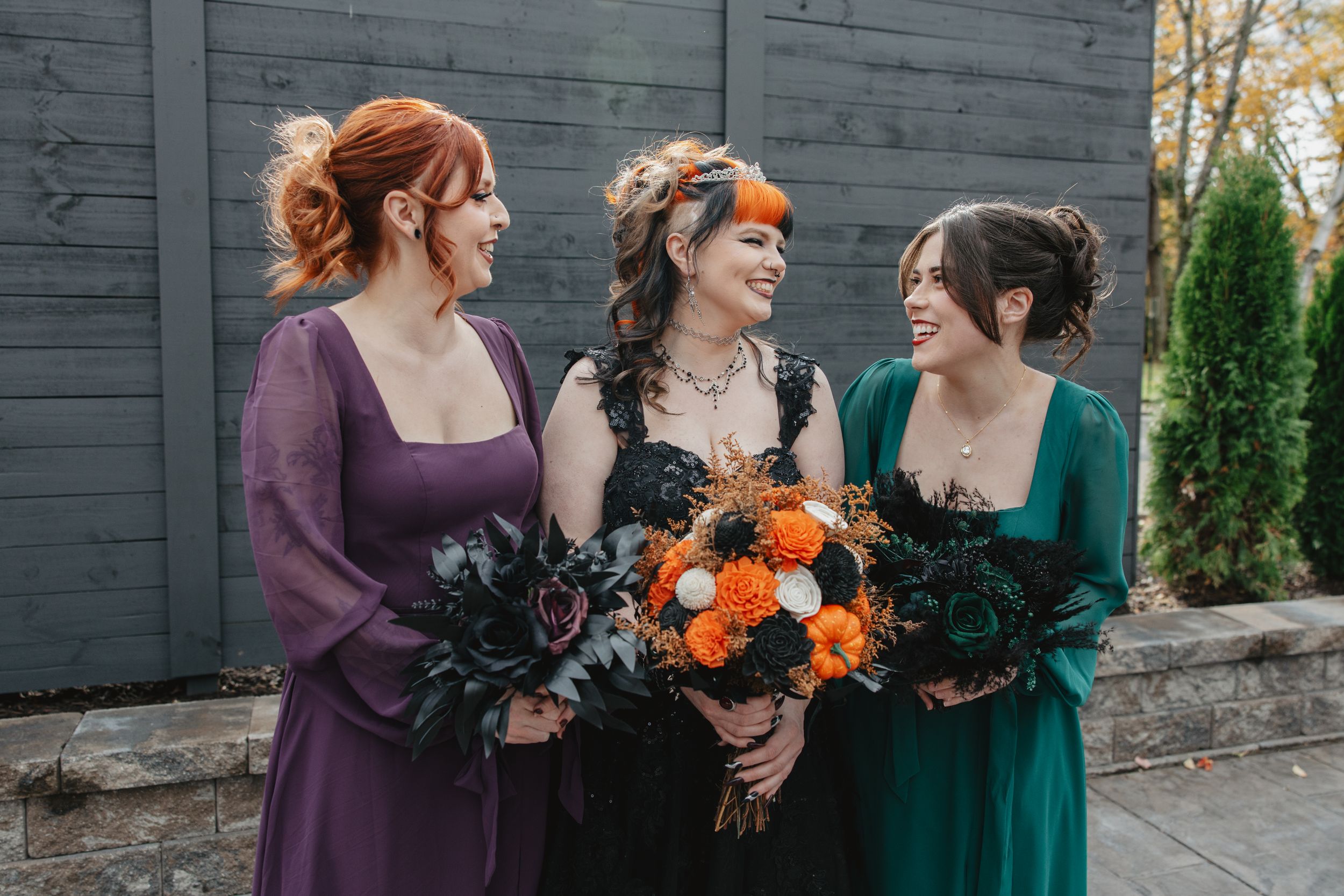 Bridesmaids in purple and emerald dresses hold orange bouquets while laughing together outdoors.