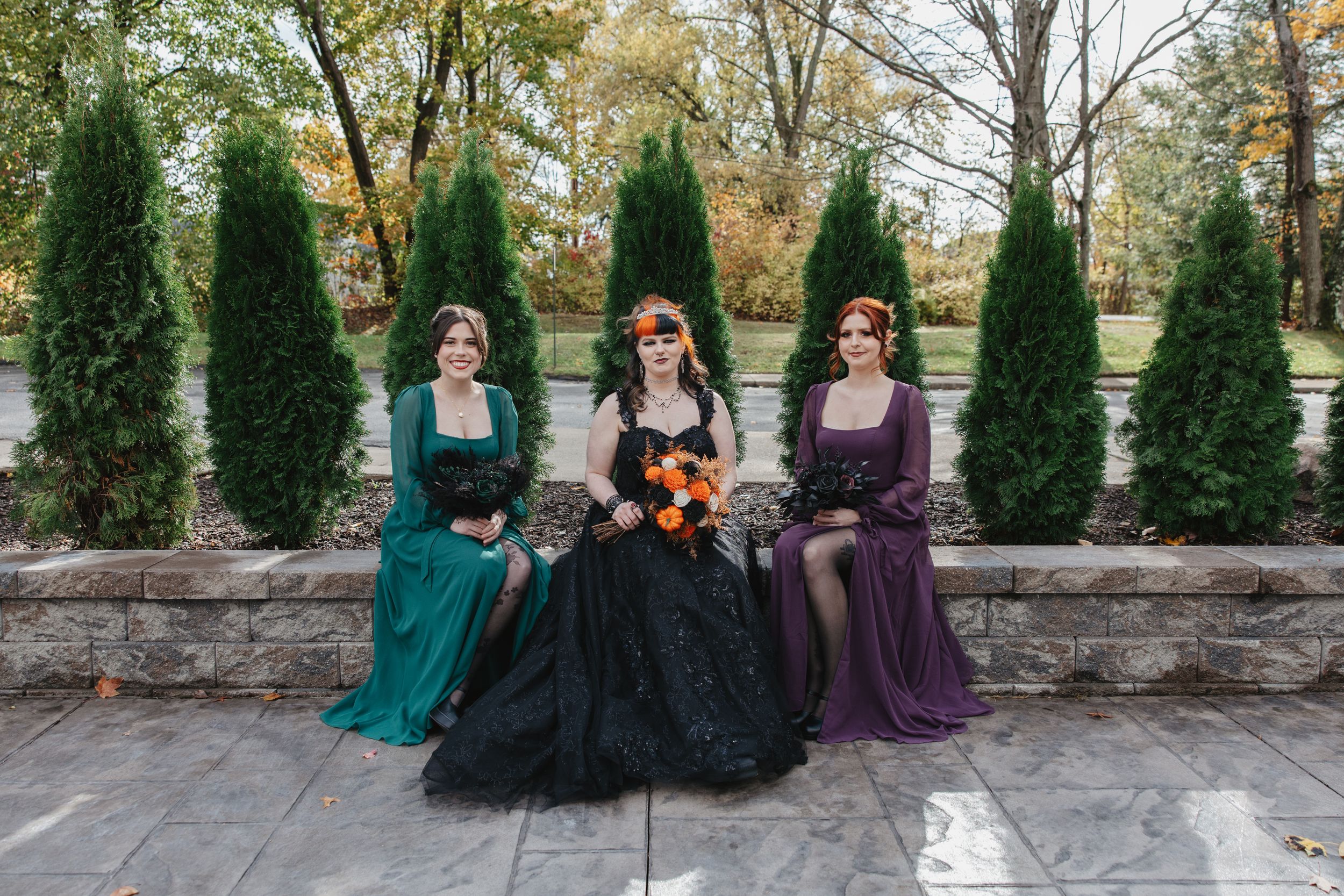 Wedding party in jewel-toned dresses poses against stone wall and evergreen trees outdoors.