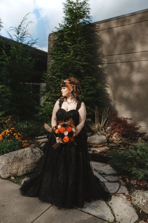 A person in a black gothic wedding dress with orange flowers poses in a garden setting with stone landscaping.