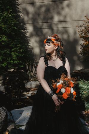 Gothic bride in black dress poses with orange flowers against moody garden backdrop.