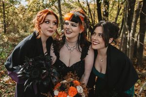 Autumn wedding party poses in forest with vibrant orange floral bouquets and black attire creating moody aesthetic.