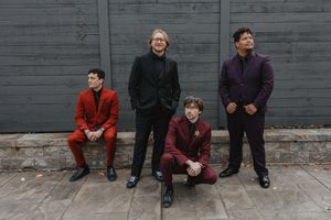 Four band members pose against a gray wall wearing formal attire in red, black and purple.