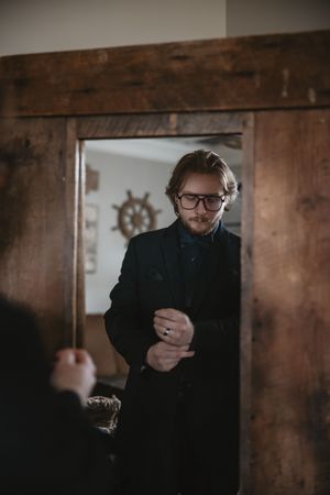 Moody portrait in a dark wooden doorway showing a figure in black coat and glasses adjusting their sleeve.
