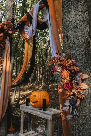Autumn wedding arch with orange and purple draping fabric, floral arrangements and carved pumpkin in forest setting.