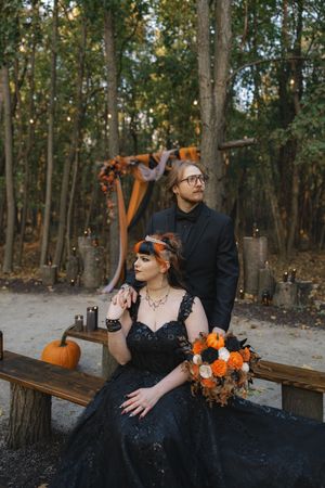 A couple in formal black attire poses on wooden benches surrounded by fall decorations and pumpkins.