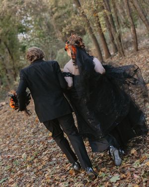 Wedding party in black attire poses dramatically on forest ground covered in fallen leaves.