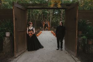 A couple stands in an outdoor garden entryway with wooden doors and autumn leaves scattered on the path.