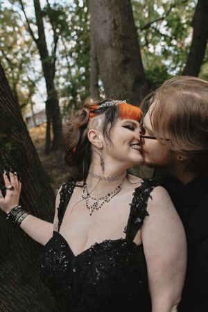 A romantic couple in gothic attire share intimate moments against a tree trunk in a woodland setting.