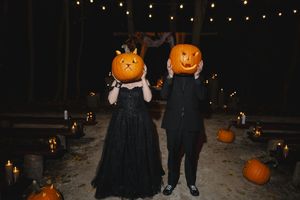 Two people wearing pumpkin heads pose together in black outfits surrounded by string lights and jack-o-lanterns at night.