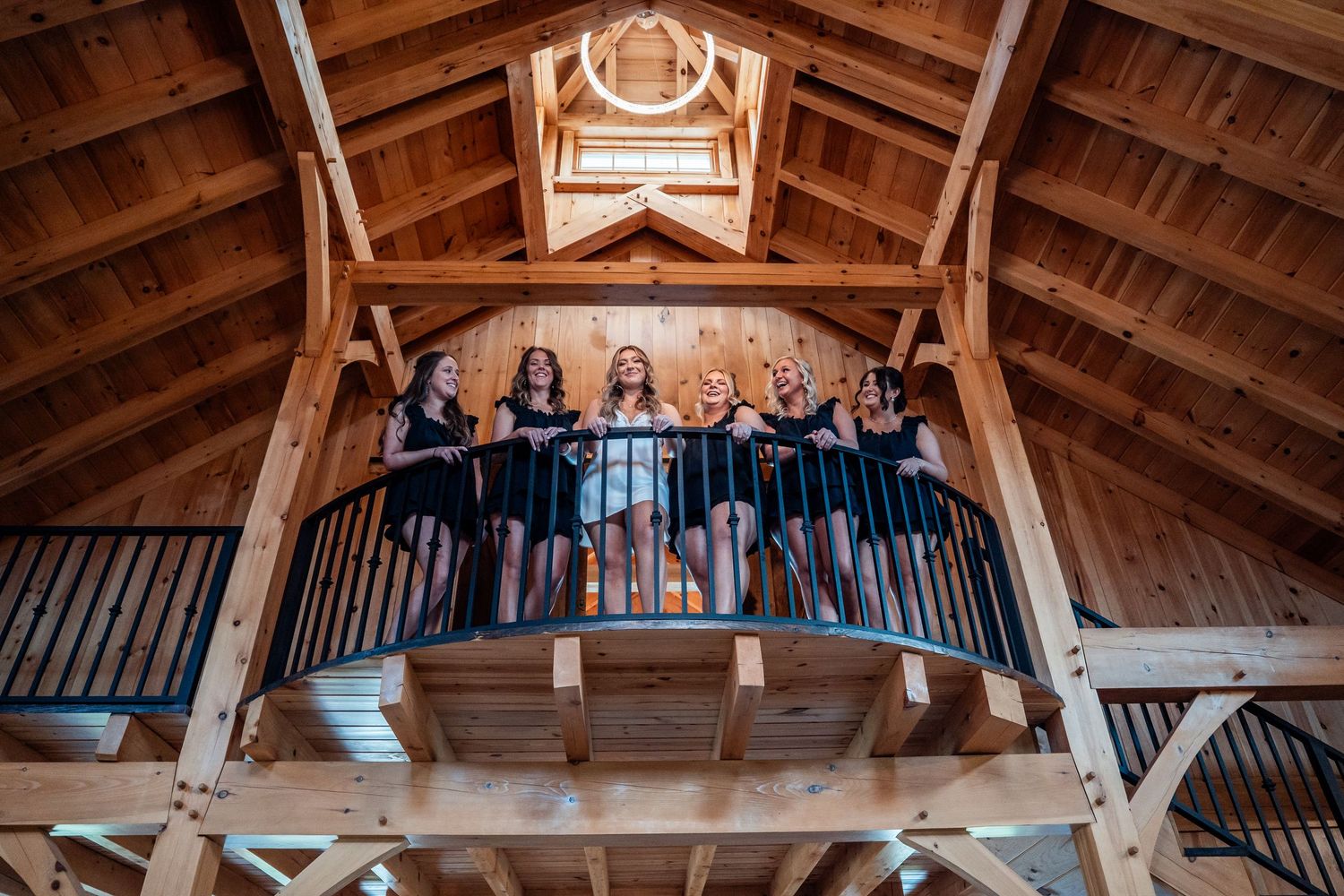 Wedding party poses on rustic wooden balcony overlooking barn venue interior.