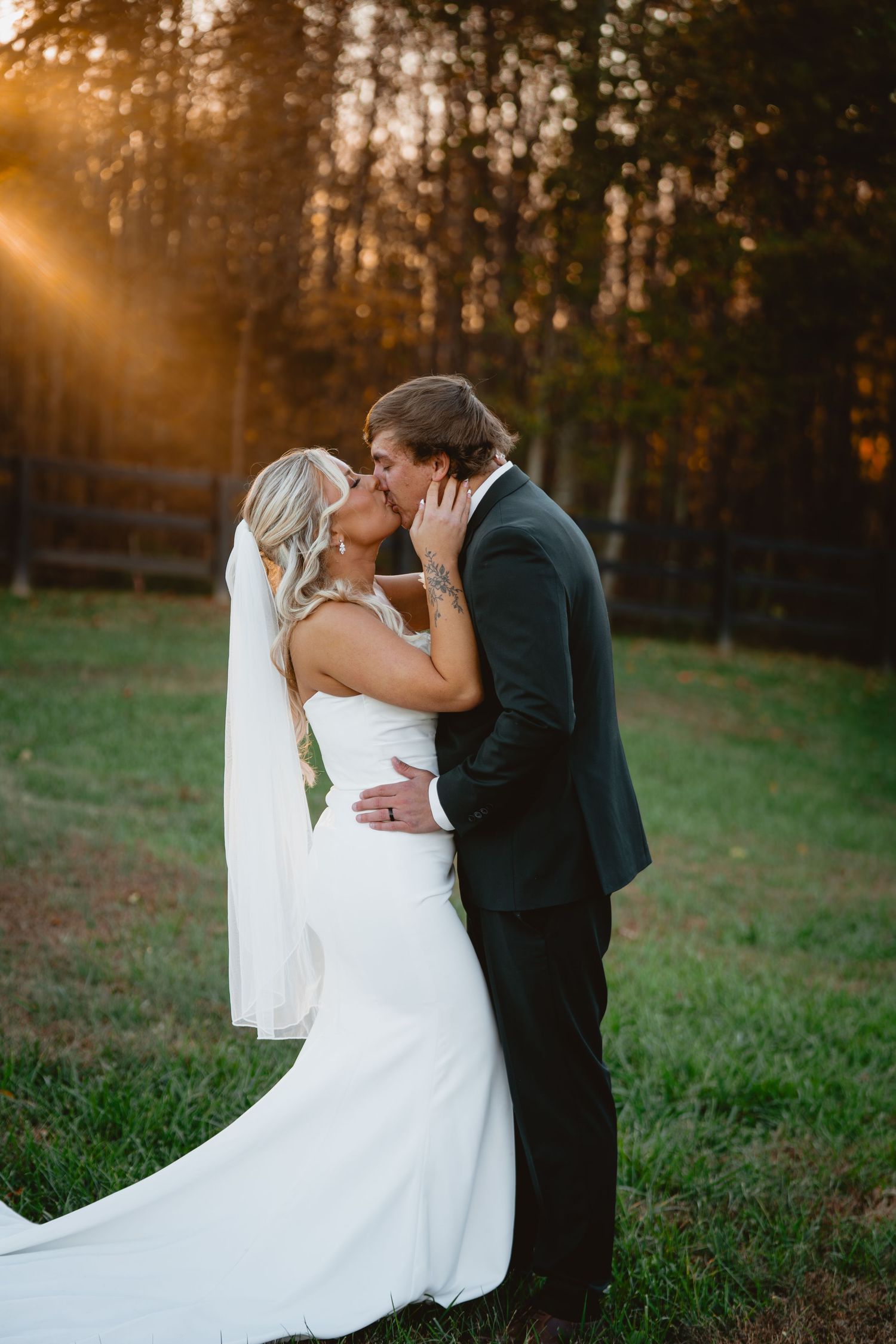 A couple shares a romantic kiss at sunset in a field with golden sunlight filtering through trees in the background.