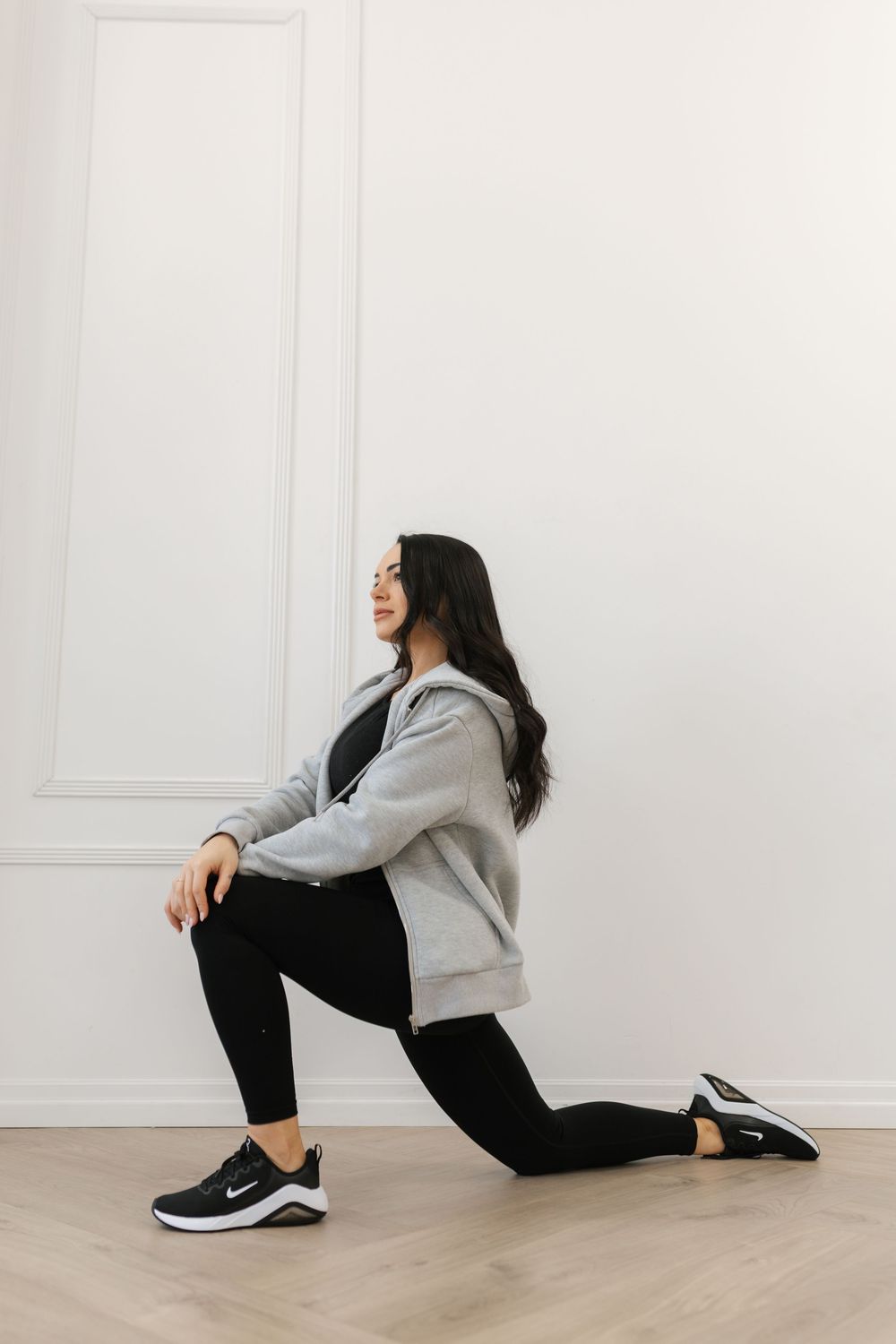 Athletic woman in gray hoodie performing a lunge exercise on hardwood floor against white wall.