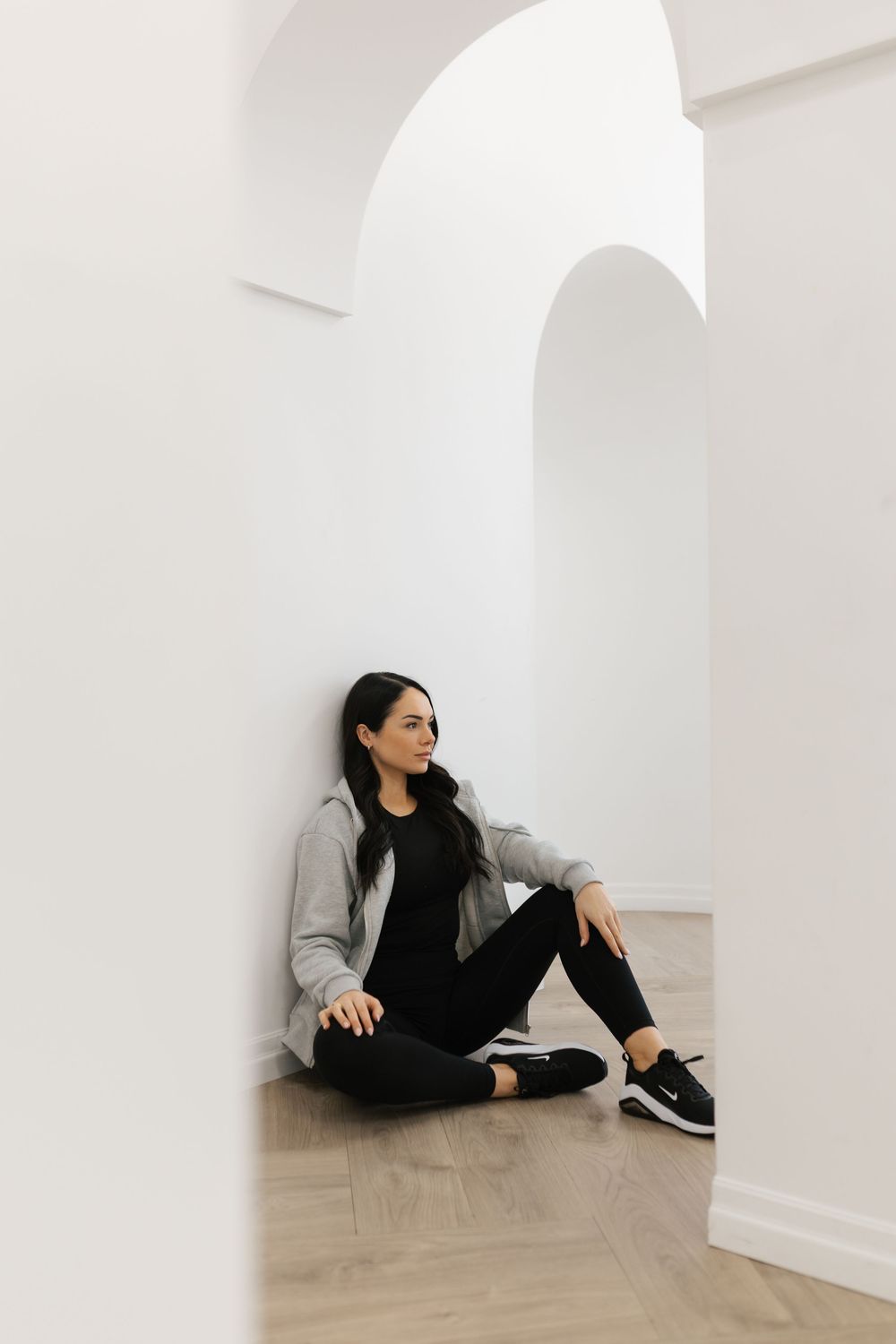 Young woman in gray hoodie sitting on hardwood floor leaning against white wall with arched doorway.
