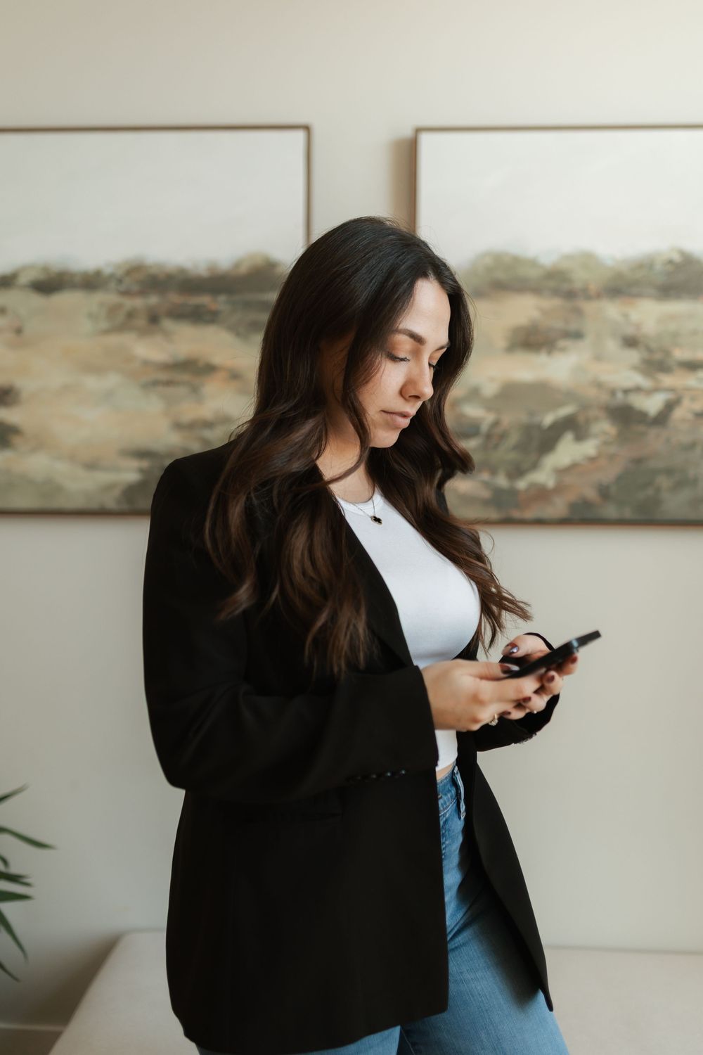 Young professional woman in black blazer and jeans checking smartphone in modern office with artwork.