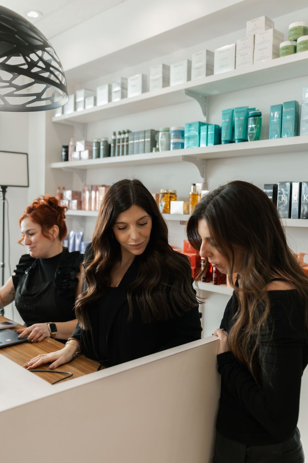 Two women in black reviewing information at a salon reception desk with product shelves behind.