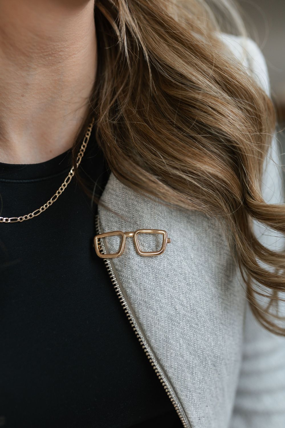 Close-up of a gold eyeglasses brooch pin on a gray blazer with a chain necklace.