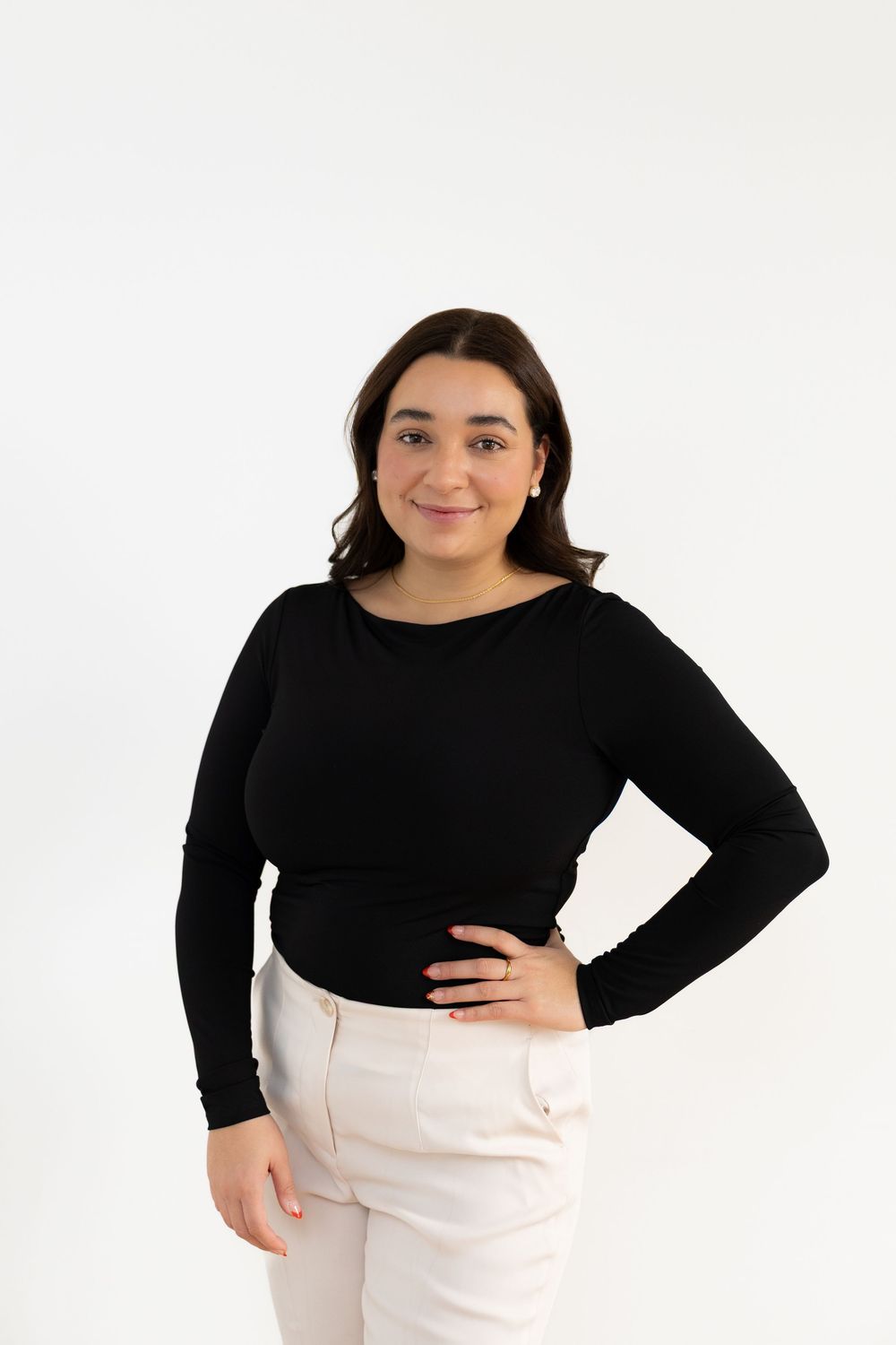 Young brunette woman in black long-sleeve top and cream pants posing against white background.