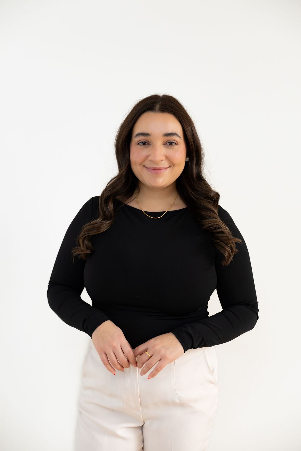 Smiling young woman in black long-sleeve top and white pants with gold necklace on white background.