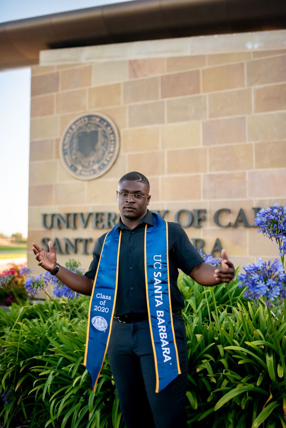 Graduate in blue sash poses in front of UCSB campus sign surrounded by purple flowers.