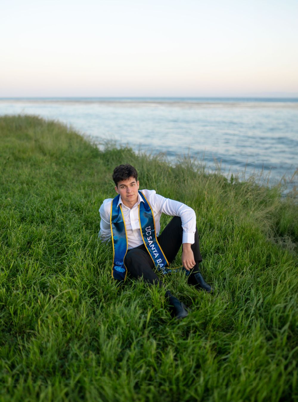 Graduate in formal attire with blue stole sits in grass overlooking ocean view.